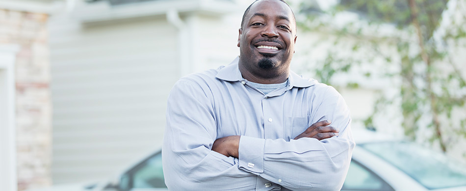 A many in a button up shirt standing in front of his home and car