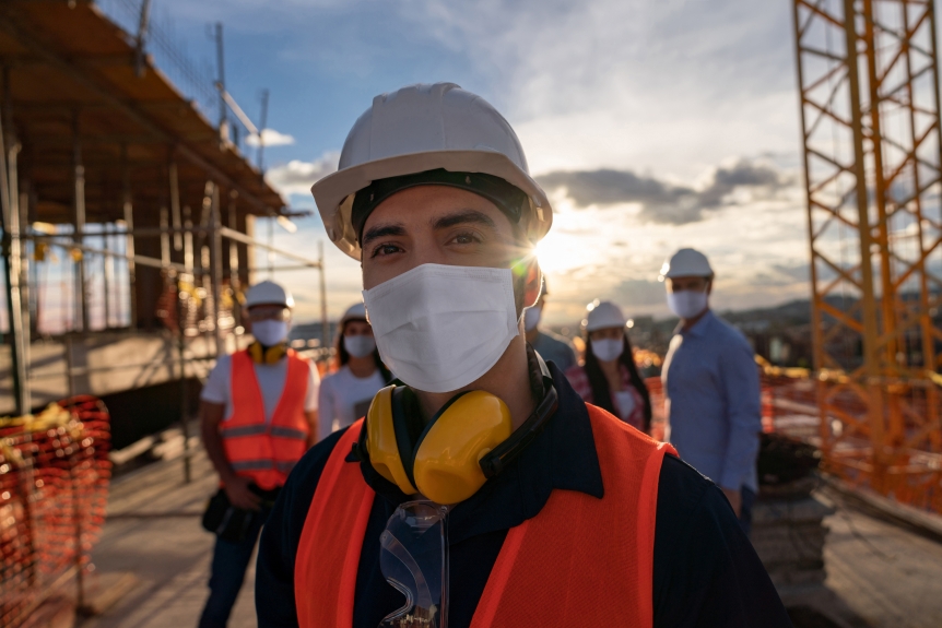 Portrait of a happy construction worker at a building site wearing a facemas with a group of workers at the background (Portrait of a happy construction worker at a building site wearing a facemas with a group of workers at the background , ASCII,